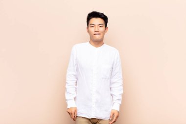 Genç Çinli adam şaşkın ve şaşkın görünüyor, gergin bir hareketle dudağını ısırıyor, düz renkli duvara karşı sorunun cevabını bilmiyor.