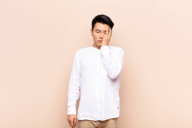 Genç Çinli bir adam, sıkıcı, sıkıcı ve sıkıcı bir işin ardından sıkılmış, hayal kırıklığına uğramış ve uykulu hisseder. Düz renkli bir duvara karşı el ele tutuşur.