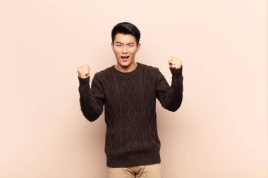 Genç Çinli adam mutlu, şaşırmış ve gururlu hissediyor, bağırıyor ve başarıyı kutluyor düz renkli duvara karşı büyük bir gülümsemeyle.