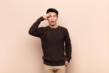 Genç Çinli adam mutlu, şaşkın ve şaşırmış görünüyor, gülümsüyor ve düz renkli duvara karşı inanılmaz ve inanılmaz iyi haberler veriyor.