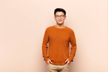 Genç Çinli adam sevinçle gülümsüyor. Olumlu, mutlu, kendinden emin ve gevşek bir ifadeyle düz duvarın karşısında.