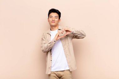 Genç Çinli adam gülümsüyor ve mutlu, sevimli, romantik ve aşık, iki elini de düz duvara dayayıp kalp şekli veriyor.