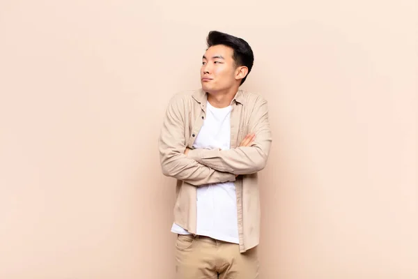 young chinese man shrugging, feeling confused and uncertain, doubting with arms crossed and puzzled look against flat color wall