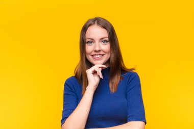 Genç kızıl saçlı kadın mutlu ve gülümsüyor, çenesi üstünde, merak ediyor ya da soru soruyor, düz duvarla seçenekleri karşılaştırıyor.