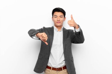 Genç Çinli bir adam kafası karışmış, bilgisiz ve kararsız hissediyor, farklı seçeneklerde iyi ve kötü şeyleri tartıyor ya da düz renkli duvarlara karşı seçenekleri tartıyor.