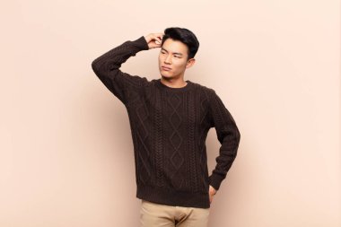 Genç Çinli adam şaşkın ve kafası karışmış hissediyor. Kafasını kaşıyor ve düz duvara bakıyor.