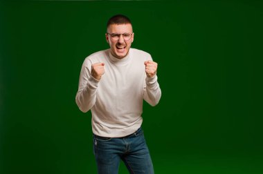 Genç yakışıklı adam sinirli, sinirli, sinirli bakışlar ve sıkı yumruklarla bağırıyor, düz arkaplana karşı öfkeli hissediyor.