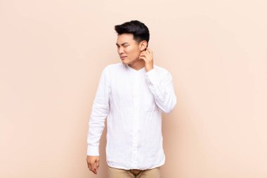 Genç Çinli bir adam stresli, hayal kırıklığına uğramış ve yorgun hissediyor, boynu ağrıyor, endişeli, sorunlu bir bakışla düz renkli duvara bakıyor.