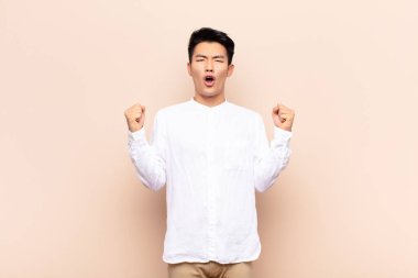 Genç Çinli bir adam sinirli bir ifadeyle ya da düz renk duvara karşı başarıyı kutlayan yumruklarla agresif bir şekilde bağırıyor.