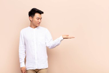 Genç Çinli bir adam kendini mutlu hissediyor ve gelişigüzel gülümsüyor. Düz renkli duvara karşı elinde tuttuğu bir nesne ya da konsept arıyor.