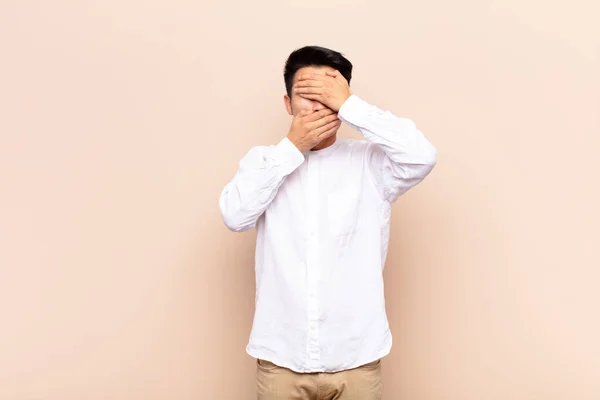 young chinese man covering face with both hands saying no to the camera! refusing pictures or forbidding photos against flat color wall