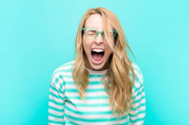 young pretty blonde woman shouting aggressively, looking very angry, frustrated, outraged or annoyed, screaming no against flat color wall clipart