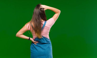 Genç, güzel bir kadın bilgisiz ve kafası karışık, bir çözüm düşünüyor, eli kalçasında, diğeri kafasında, yeşil arka planda.