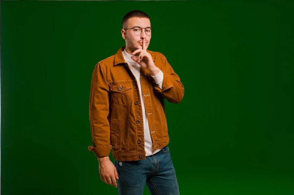young handsome man asking for silence and quiet, gesturing with finger in front of mouth, saying shh or keeping a secret against flat background