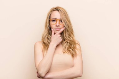 Genç, güzel sarışın kadın ciddi, düşünceli ve güvensiz görünüyor, bir kolu çapraz ve çene üzerinde, düz renkli duvarla seçenekleri tartıyor