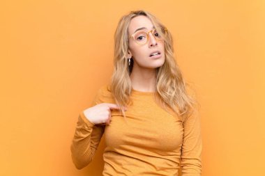 Genç, güzel, sarışın bir kadın şaşkın, şaşkın ve güvensiz hissediyor. Kendini gösterip kim olduğumu soruyor, ben mi? yassı renk duvarına karşı