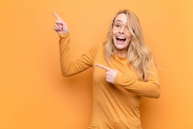 Genç, güzel, sarışın bir kadın sevinçli ve şaşırmış hissediyor. Şok olmuş bir ifadeyle gülümsüyor ve düz renkli duvara işaret ediyor.