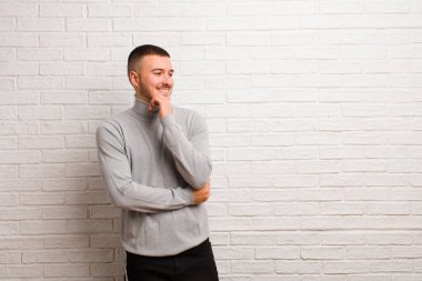 Genç yakışıklı adam mutlu, kendinden emin bir ifadeyle gülümsüyor, elleri çenesinde, merak ediyor ve düz arkaplana bakıyor.