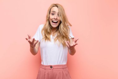 Genç ve güzel sarışın kadın ağzı açık ve şaşırmış, şok olmuş ve şaşırmış bir şekilde düz renkli duvara karşı inanılmaz bir sürprizle