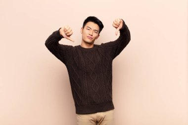 Genç Çinli adam üzgün, hayal kırıklığına uğramış ya da kızgın görünüyor. Anlaşmazlıkta baş parmağını gösteriyor. Düz renkli duvara karşı hayal kırıklığına uğramış hissediyor.