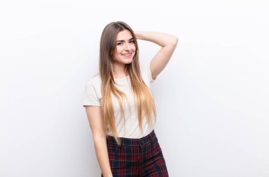 Genç, güzel bir kadın neşeli ve tesadüfen gülümsüyor, beyaz duvara karşı pozitif, mutlu ve kendinden emin bir bakışla göğüs göğüse tutuşuyor.