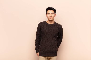 Genç Çinli adam gururlu, kendinden emin, havalı, küstah ve küstah görünüyor, gülümsüyor, düz renkli duvara karşı başarılı hissediyor.