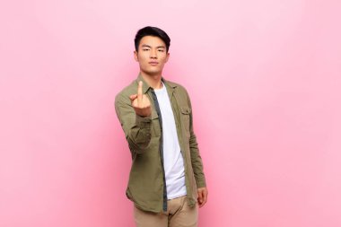 Genç Çinli bir adam kızgın, sinirli, isyankar ve agresif orta parmağını sallayarak düz renkli duvara karşı savaşıyor.