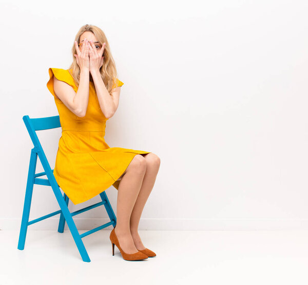 young pretty blonde woman covering face with hands, peeking between fingers with surprised expression and looking to the side against flat color wall