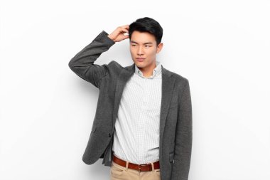 Genç Çinli adam şaşkın ve kafası karışmış hissediyor. Kafasını kaşıyor ve düz duvara bakıyor.