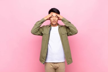 Genç Çinli adam konsantre, düşünceli ve ilham verici görünüyor, beyin fırtınası yapıyor ve düz renk duvarına karşı elleri alnında hayal ediyor.