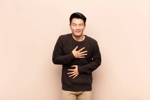 young chinese man laughing out loud at some hilarious joke, feeling happy and cheerful, having fun against flat color wall