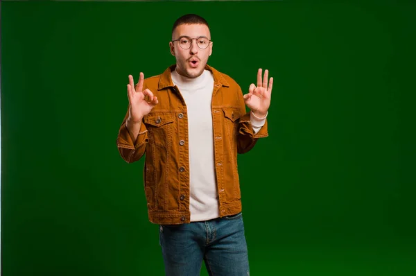 stock image young handsome man feeling shocked, amazed and surprised, showing approval making okay sign with both hands against flat background