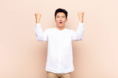 Genç Çinli bir adam inanılmaz bir başarıyı kutluyor. Kazanan gibi. Heyecanlı ve mutlu görünüyor. yassı renk duvarına karşı