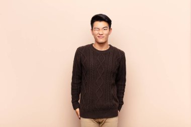 Genç Çinli adam mutlu ve arkadaş canlısı görünüyor, gülümsüyor ve düz renkli duvara karşı pozitif bir tavırla sana göz kırpıyor.