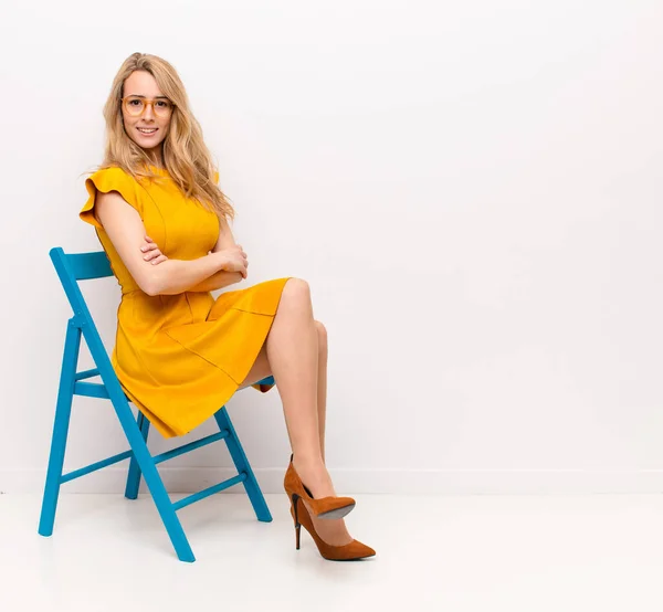 young pretty blonde woman smiling to camera with crossed arms and a happy, confident, satisfied expression, lateral view against flat color wall