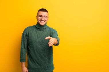 Genç yakışıklı adam gülümsüyor, mutlu, kendinden emin ve arkadaş canlısı görünüyor. Anlaşmayı bağlamak için el sıkışmayı teklif ediyor. Düz arkaplan karşısında işbirliği yapıyor.