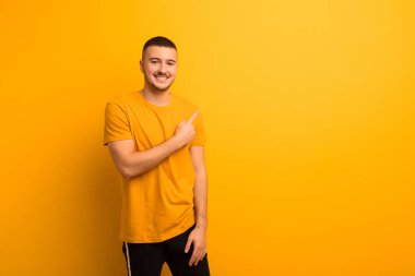 Genç yakışıklı adam neşeyle gülümsüyor, mutlu hissediyor ve yan ve yukarı doğru işaret ediyor, fotokopi alanındaki nesneleri düz arkaplana karşı gösteriyor.