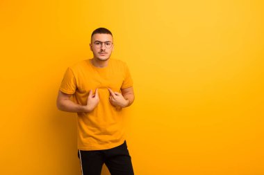 Genç yakışıklı adam, kafası karışmış ve tuhaf bakışlarıyla kendini işaret ediyor. Düz arkaplan karşısında seçildiği için şaşırmış ve şaşırmış.