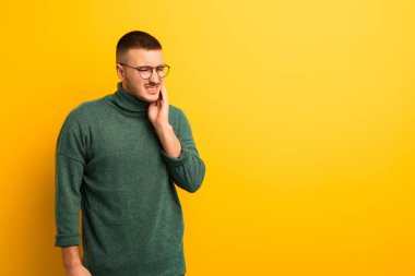 Genç yakışıklı adam yanağını tutuyor ve acı çekiyor, hasta, sefil ve mutsuz hissediyor, düz arkaplanda bir dişçi arıyor.