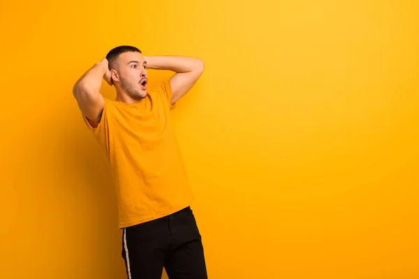 young handsome man with open mouth, looking horrified and shocked because of a terrible mistake, raising hands to head against flat background