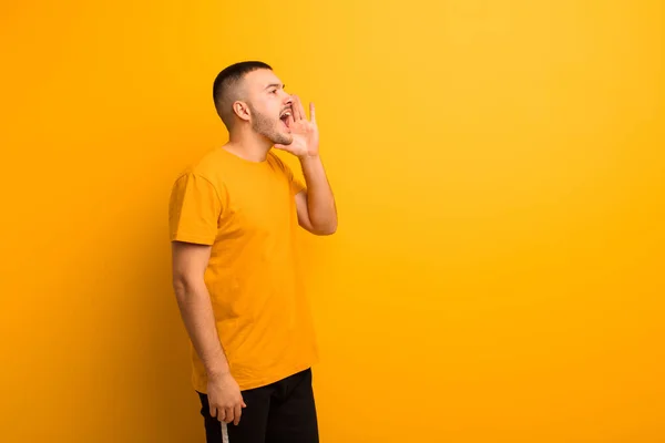young handsome man profile view, looking happy and excited, shouting and calling to copy space on the side against flat background