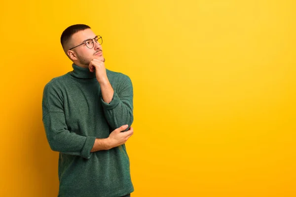 young handsome man feeling thoughtful, wondering or imagining ideas, daydreaming and looking up to copy space against flat background