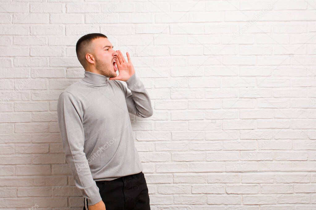 young handsome man yelling loudly and angrily to copy space on the side, with hand next to mouth against flat wall
