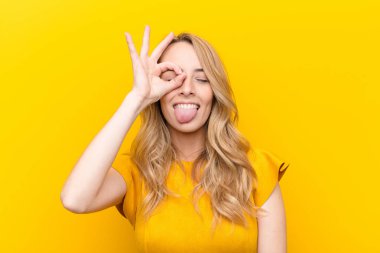 young pretty blonde woman smiling happily with funny face, joking and looking through peephole, spying on secrets against flat color wall clipart