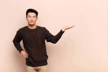 Genç Çinli adam gülümsüyor, kendinden emin, başarılı ve mutlu hissediyor, fotokopi odasında düz renkli duvara karşı konsept veya fikir gösteriyor.