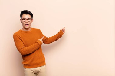 Genç Çinli adam şok olmuş ve şaşırmış hissediyor, yan taraftaki boşluğu işaret ederek, düz renkli duvara karşı şaşkın, açık ağızlı bir bakışla