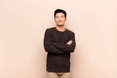 Genç Çinli adam kameraya çapraz kollarla gülümsüyor ve mutlu, kendinden emin, tatmin olmuş bir ifade, düz renkli duvara karşı yan bakış.