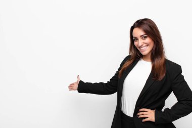 young pretty businesswoman feeling happy and cheerful, smiling and welcoming you, inviting you in with a friendly gesture against white wall clipart