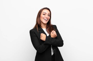 Genç ve güzel iş kadını neşeli bir şekilde gülümsüyor ve kollarını kavuşturup kollarını kavuşturup beyaz duvara karşı mutlu ve pozitif hissediyor.