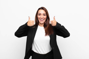 young pretty businesswoman smiling broadly looking happy, positive, confident and successful, with both thumbs up against white wall clipart